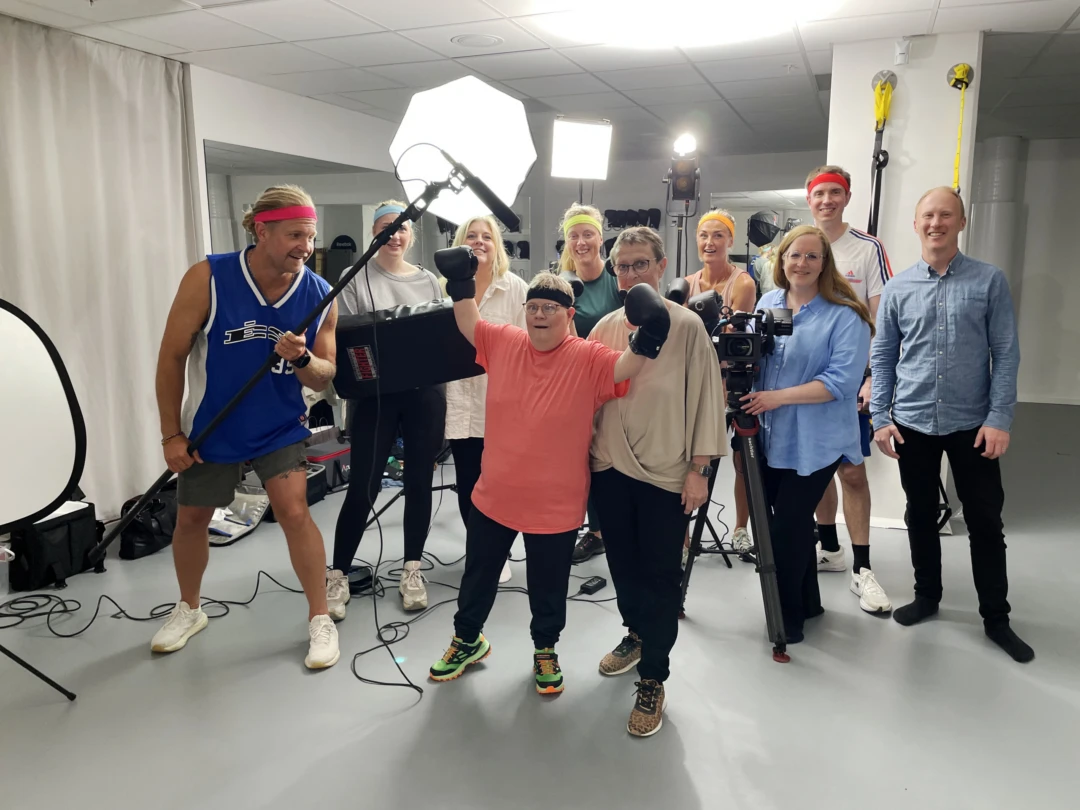 En grupp människor står uppställda i en träningslokal. De har filmkamera, mikrofonbom och flera studiolampor i bakgrunden. Eva och Lena står i mitten.