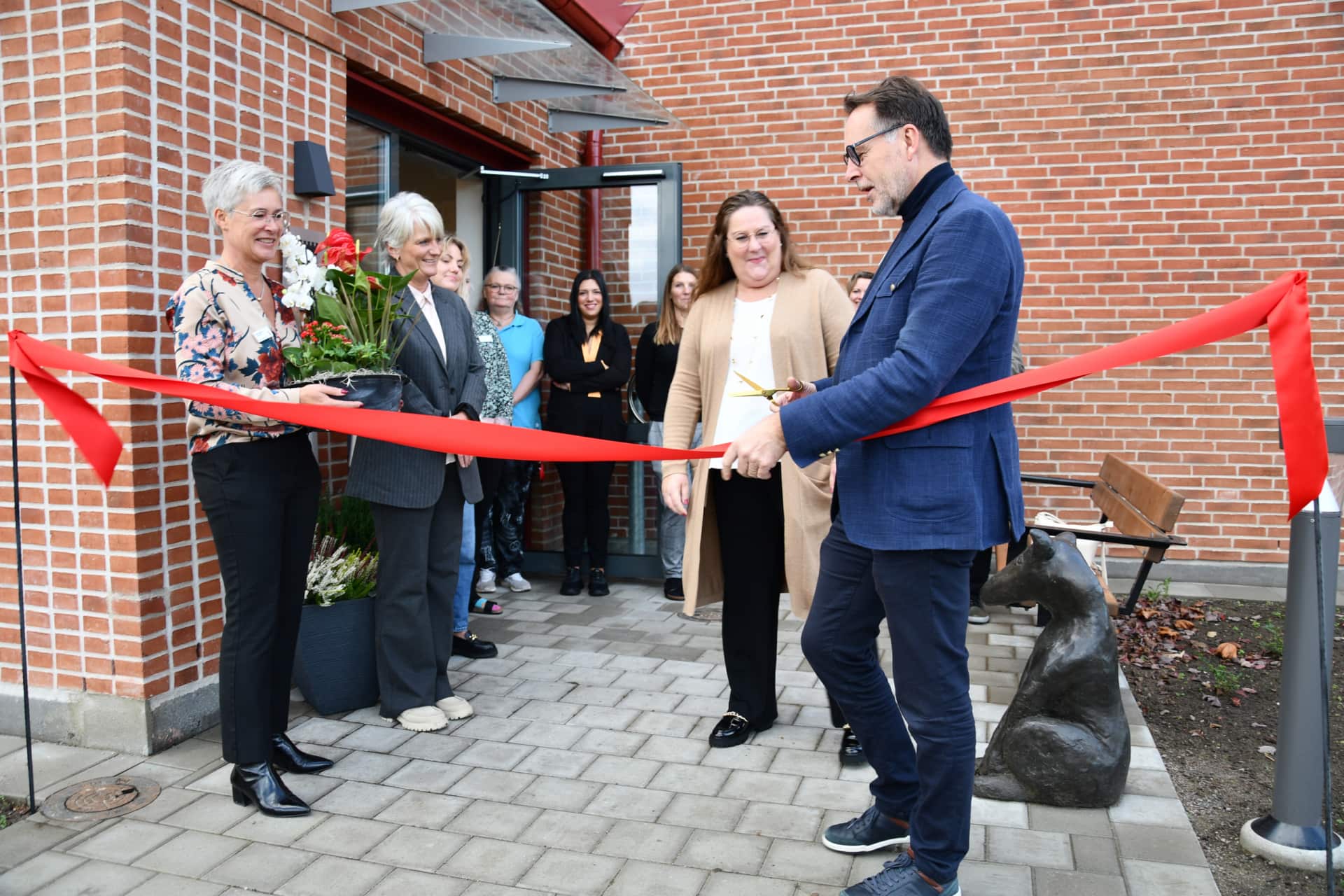 Ett rött band på väg att klippas framför hus med förväntansfulla personer.