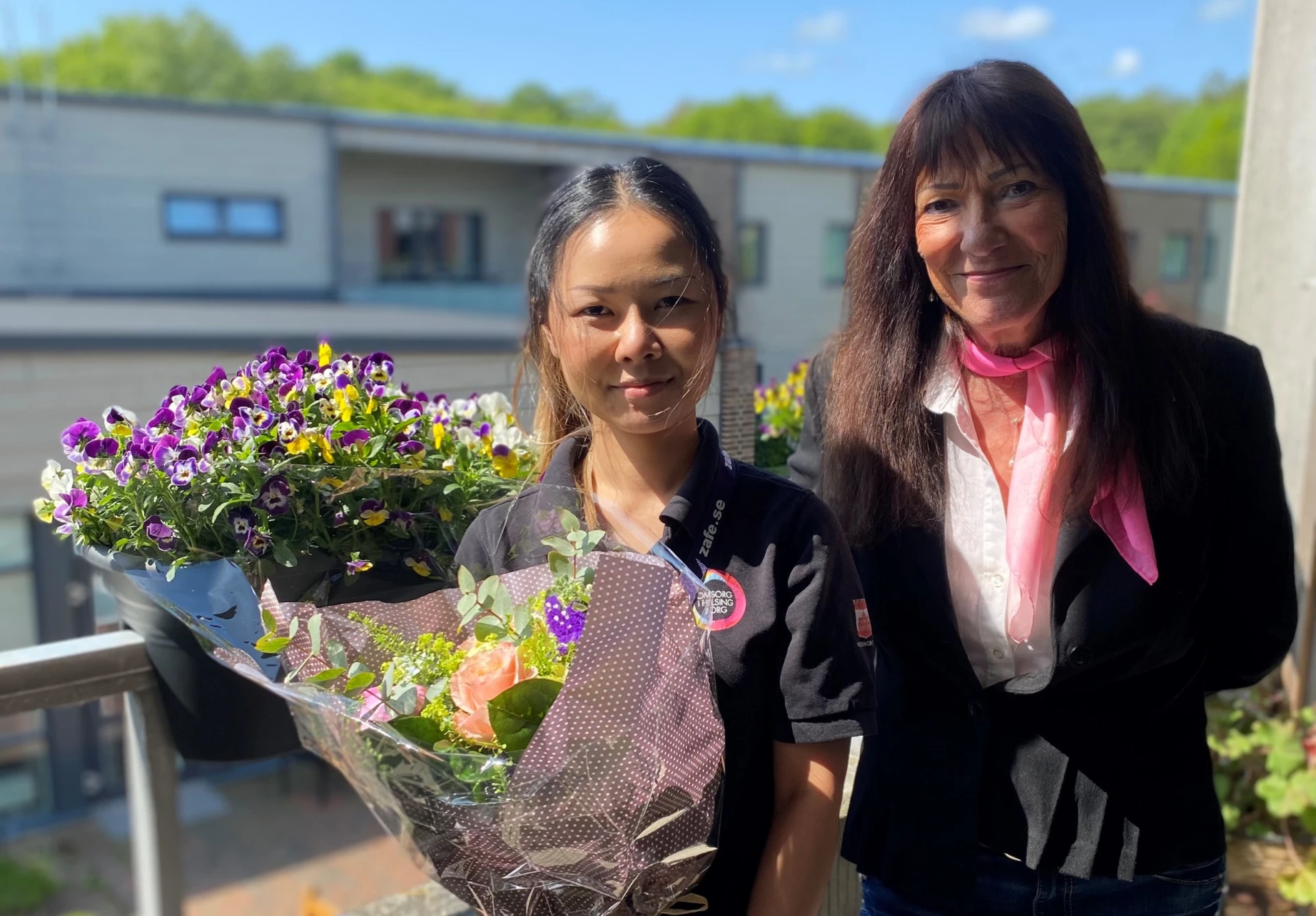 två kvinnor med mörkt långt hår, står på en balkong. Kvinnan till vänster håller i en stor blombukett