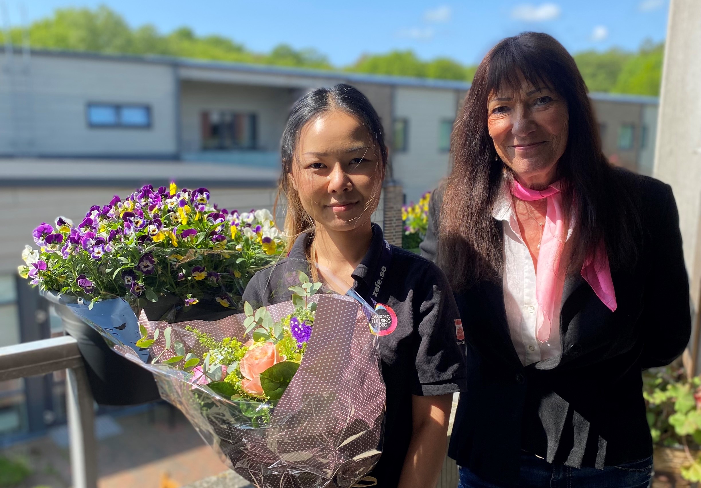 två kvinnor med mörkt långt hår, står på en balkong. Kvinnan till vänster håller i en stor blombukett
