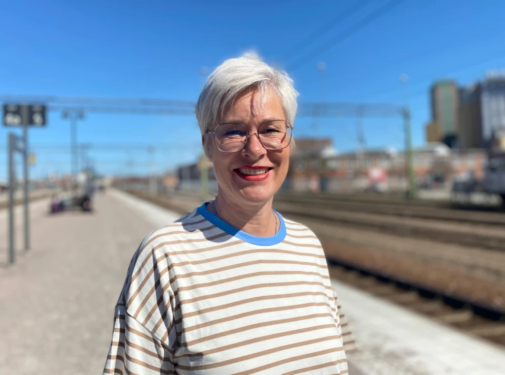 Kvinna står utomhus vid järnvägsspåret och ler i solen. Hon har kort ljust hår och glasögon.