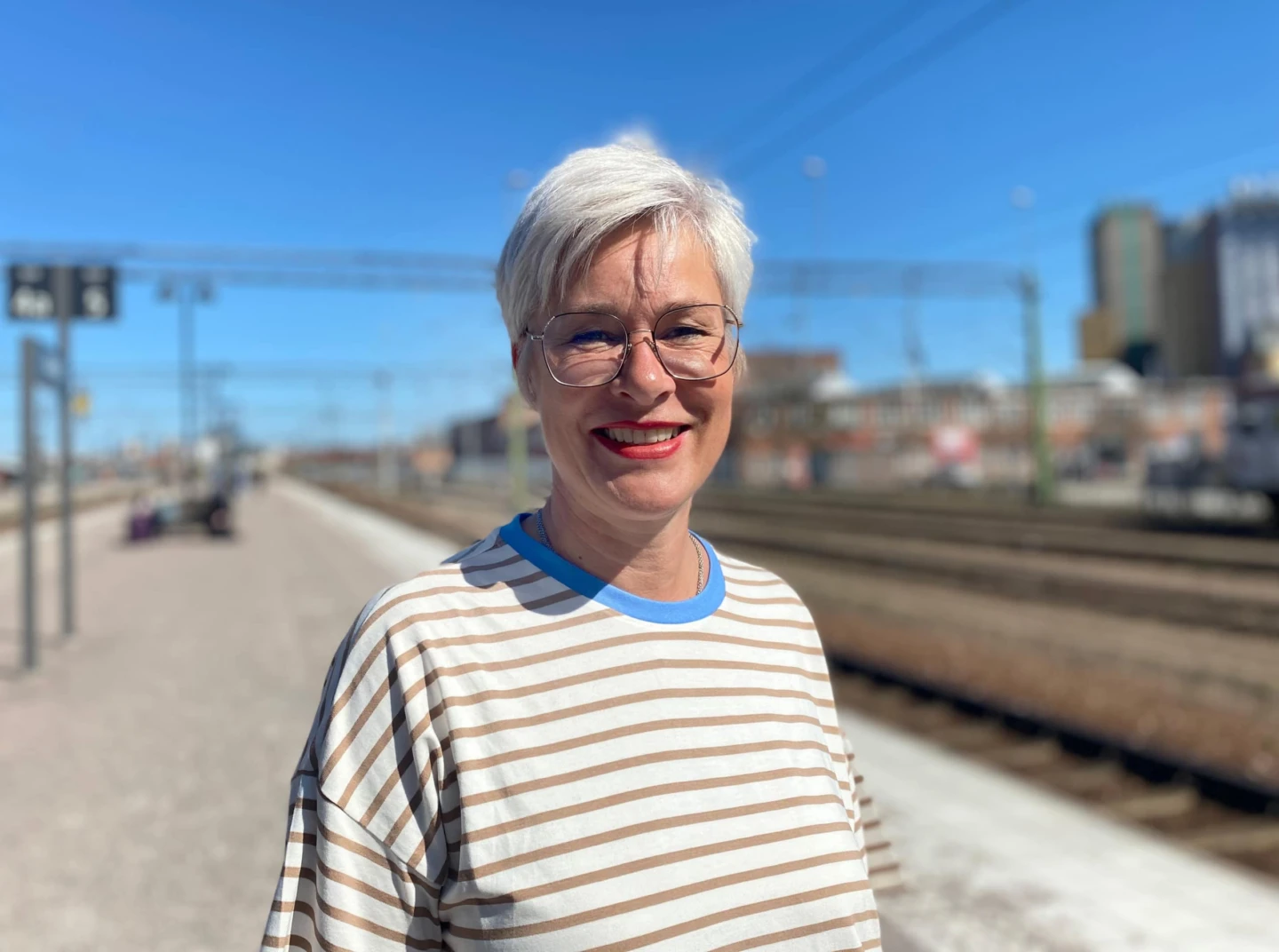 Kvinna står utomhus vid järnvägsspåret och ler i solen. Hon har kort ljust hår och glasögon.