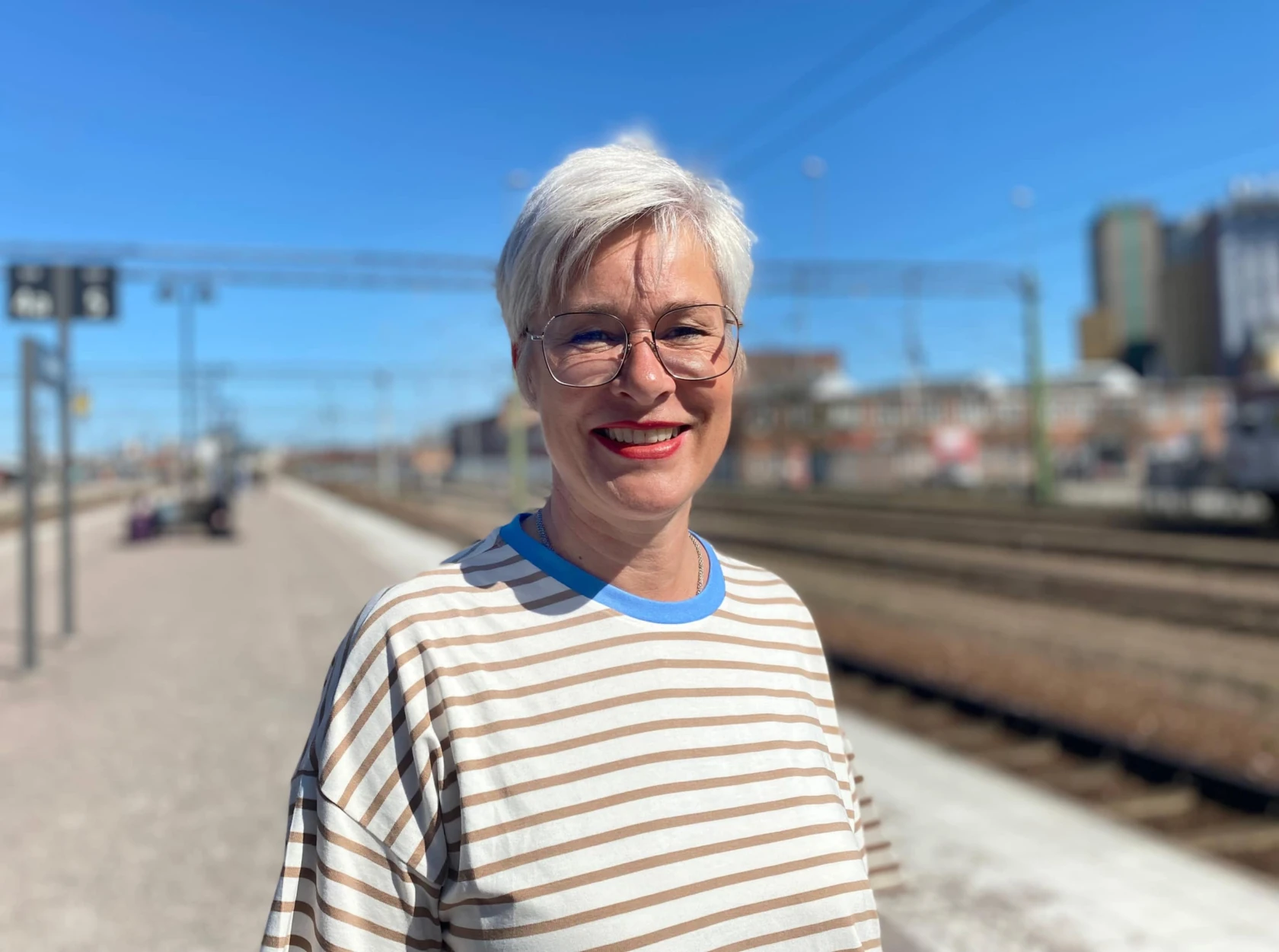 Kvinna står utomhus vid järnvägsspåret och ler i solen. Hon har kort ljust hår och glasögon.