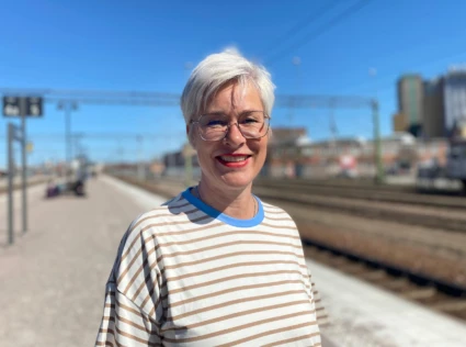 Kvinna står utomhus vid järnvägsspåret och ler i solen. Hon har kort ljust hår och glasögon.