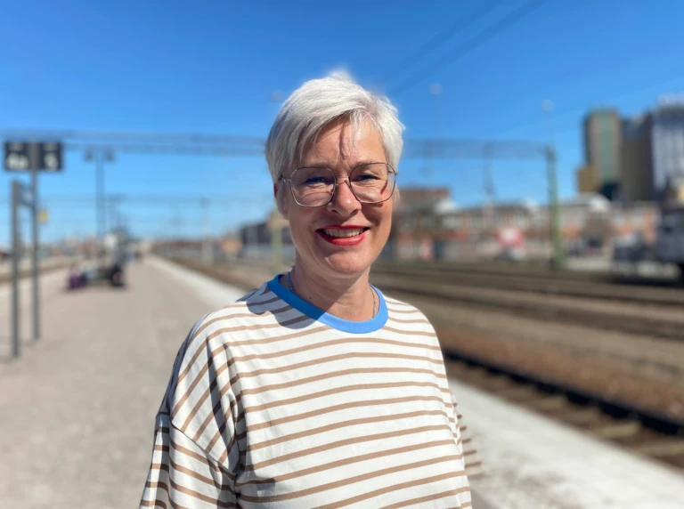 Kvinna står utomhus vid järnvägsspåret och ler i solen. Hon har kort ljust hår och glasögon.