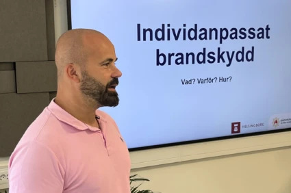 Man med skägg i rosa tröja står framför en datorskärm med en presentation.