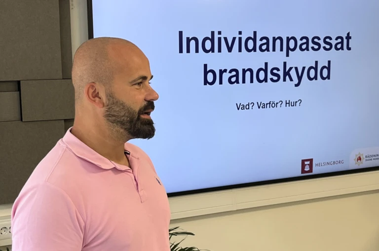 Man med skägg i rosa tröja står framför en datorskärm med en presentation.