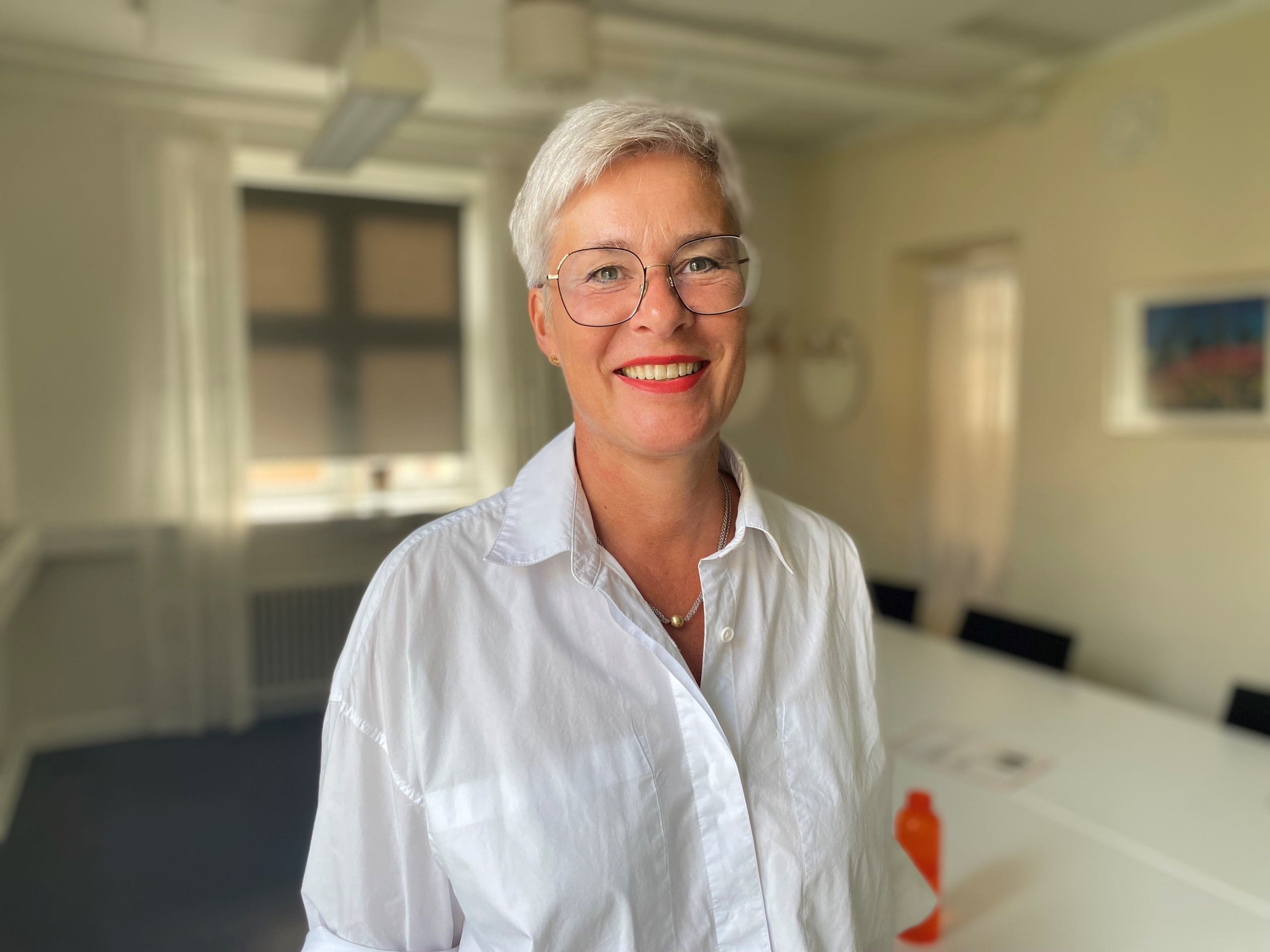 Kvinna med vit skjorta, glasögon och kort hår tittar in i kameran och ler
