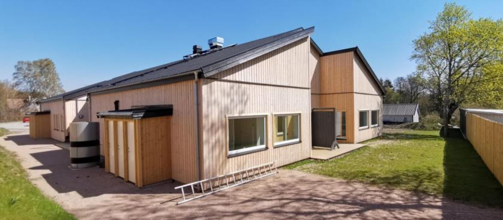 Ett enplanshus med ljusbrun fasad. grönska runtomkring. Himlen är blå.