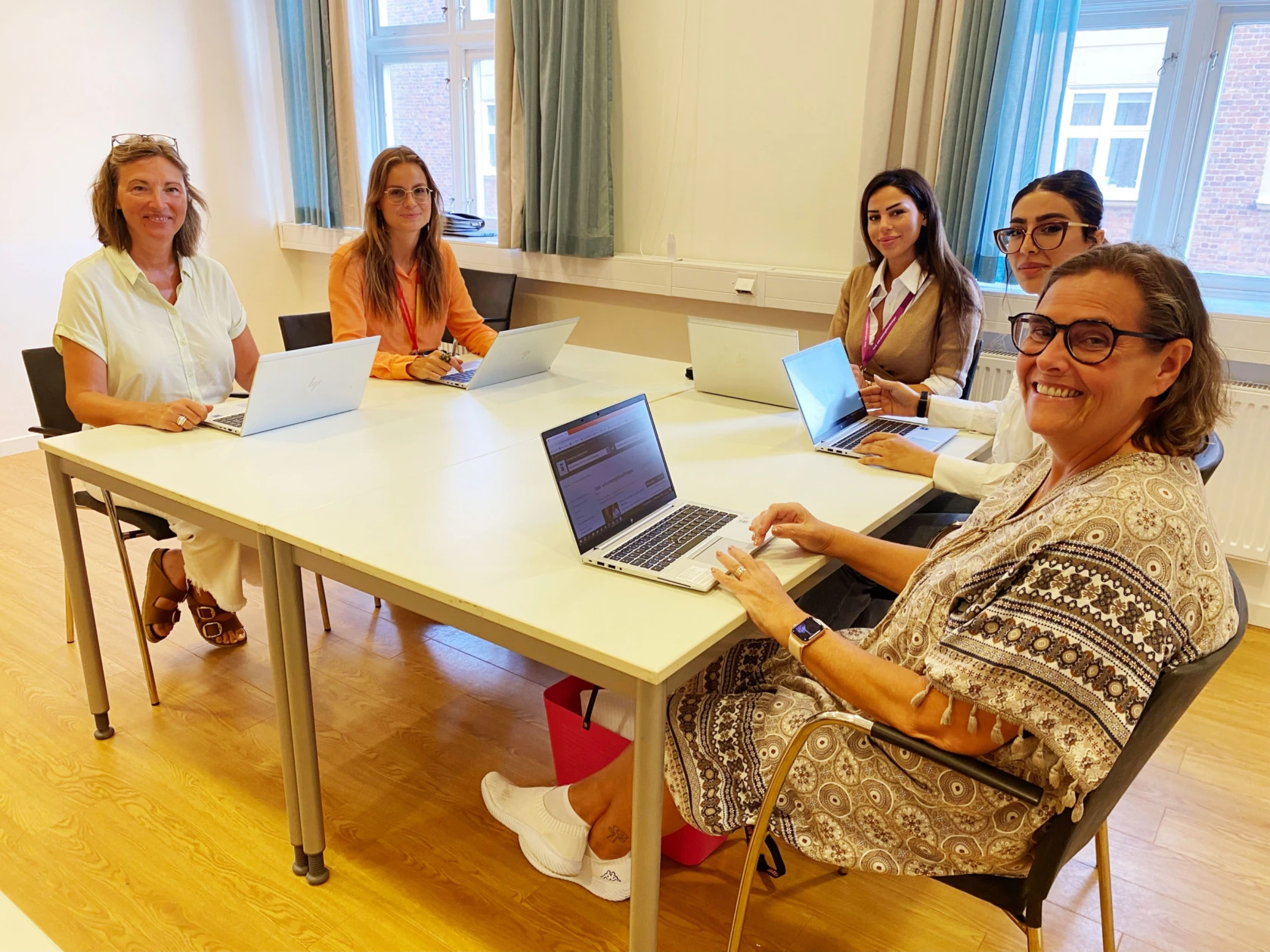 Sex kvinnor sitter runt ett bord med datorer tittar in i kameran och ler