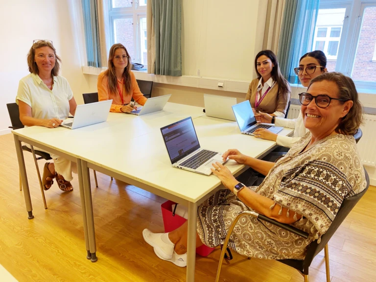 Sex kvinnor sitter runt ett bord med datorer tittar in i kameran och ler