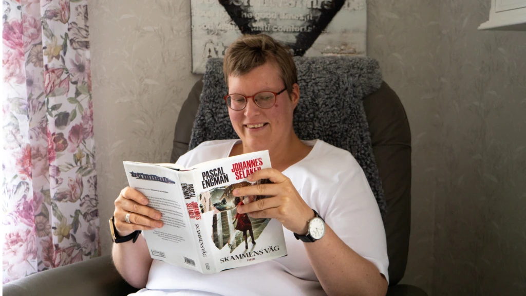 Kvinna i glasögon och kort mörkt hår sitter i en fåtölj och läser en bok.