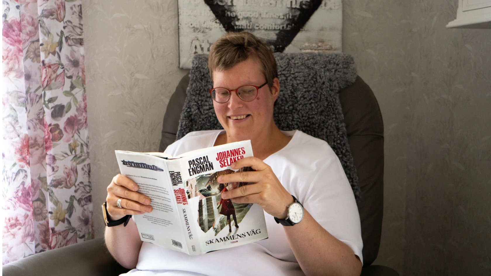 Kvinna i glasögon och kort mörkt hår sitter i en fåtölj och läser en bok.