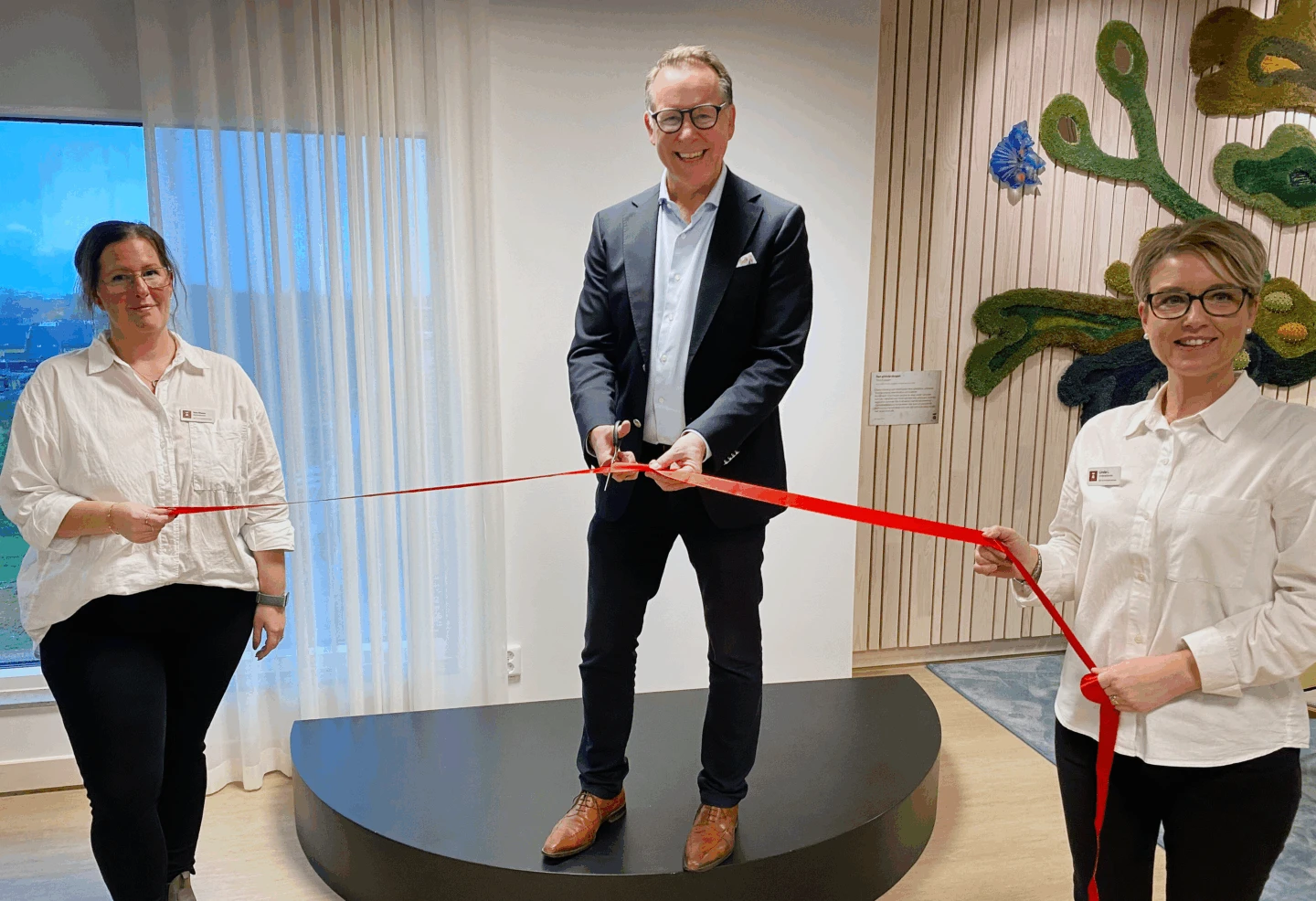 Man står på podium, håller i en sax på väg att klippa ett rött band. Bredvid står två kvinnor på varsin sida och håller i bandet.