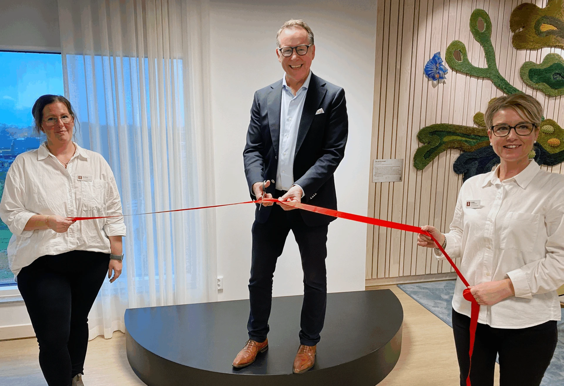 Man står på podium, håller i en sax på väg att klippa ett rött band. Bredvid står två kvinnor på varsin sida och håller i bandet.
