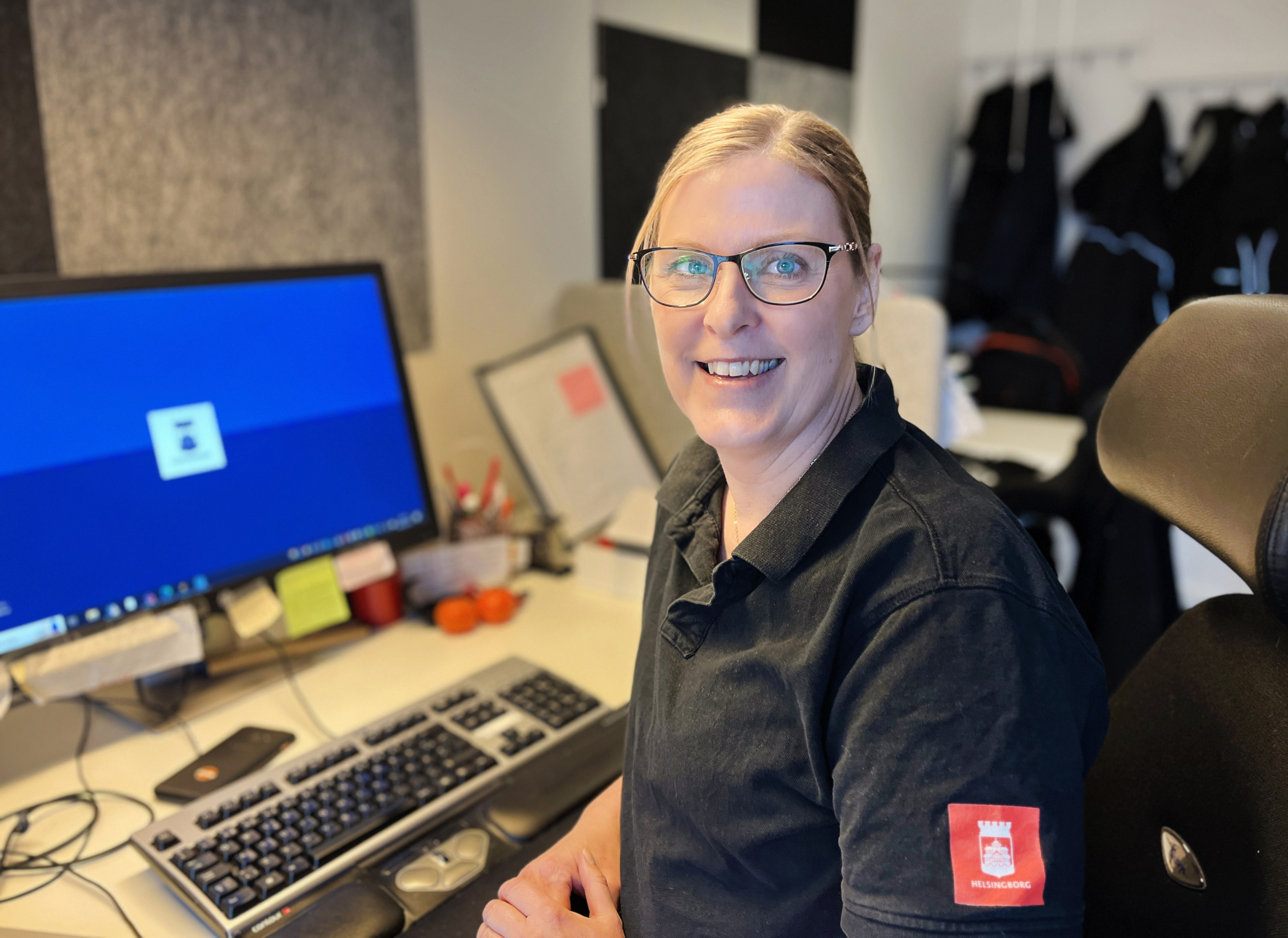 En kvinna sitter framför en datorskärm, ler och tittar in i kameran.