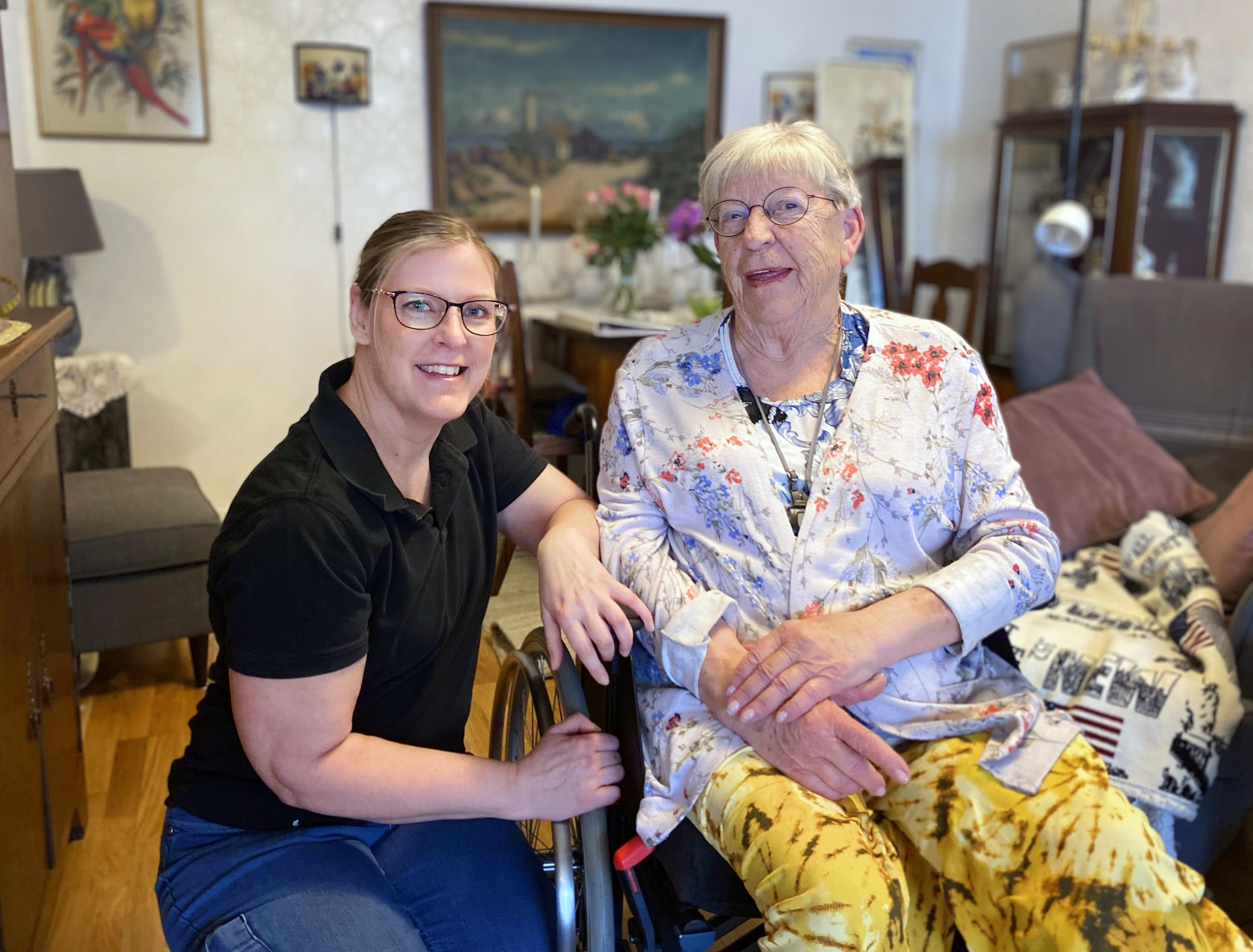 En yngre kvinna sitter på huk bredvid en äldre kvinna i rullstol. Båda tittar in i kameran.