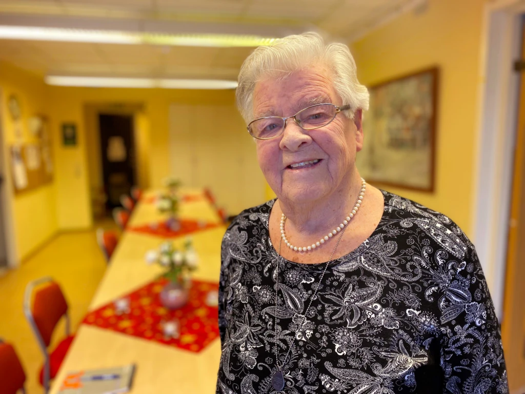 Äldre kvinna med vitt hår, står framför ett bord och ler.