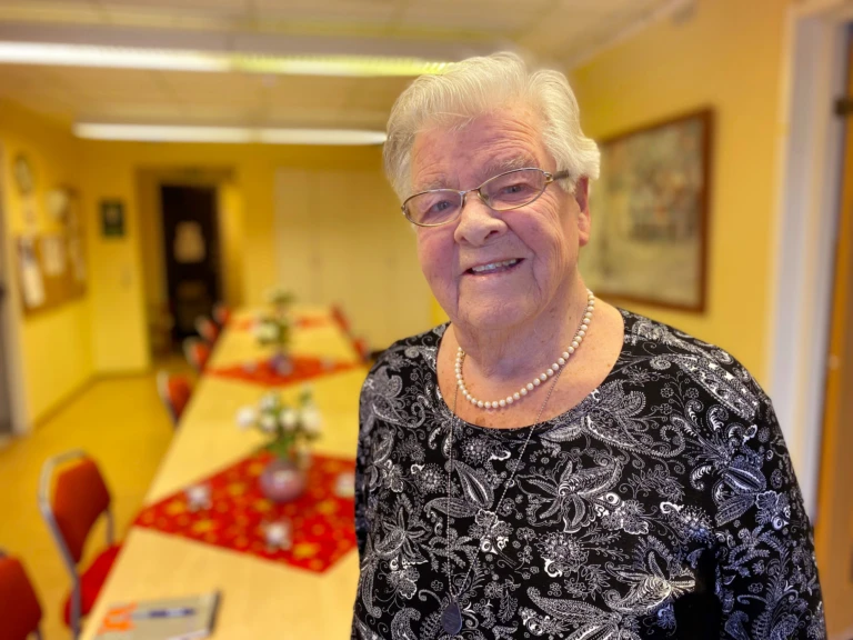 Äldre kvinna med vitt hår, står framför ett bord och ler.