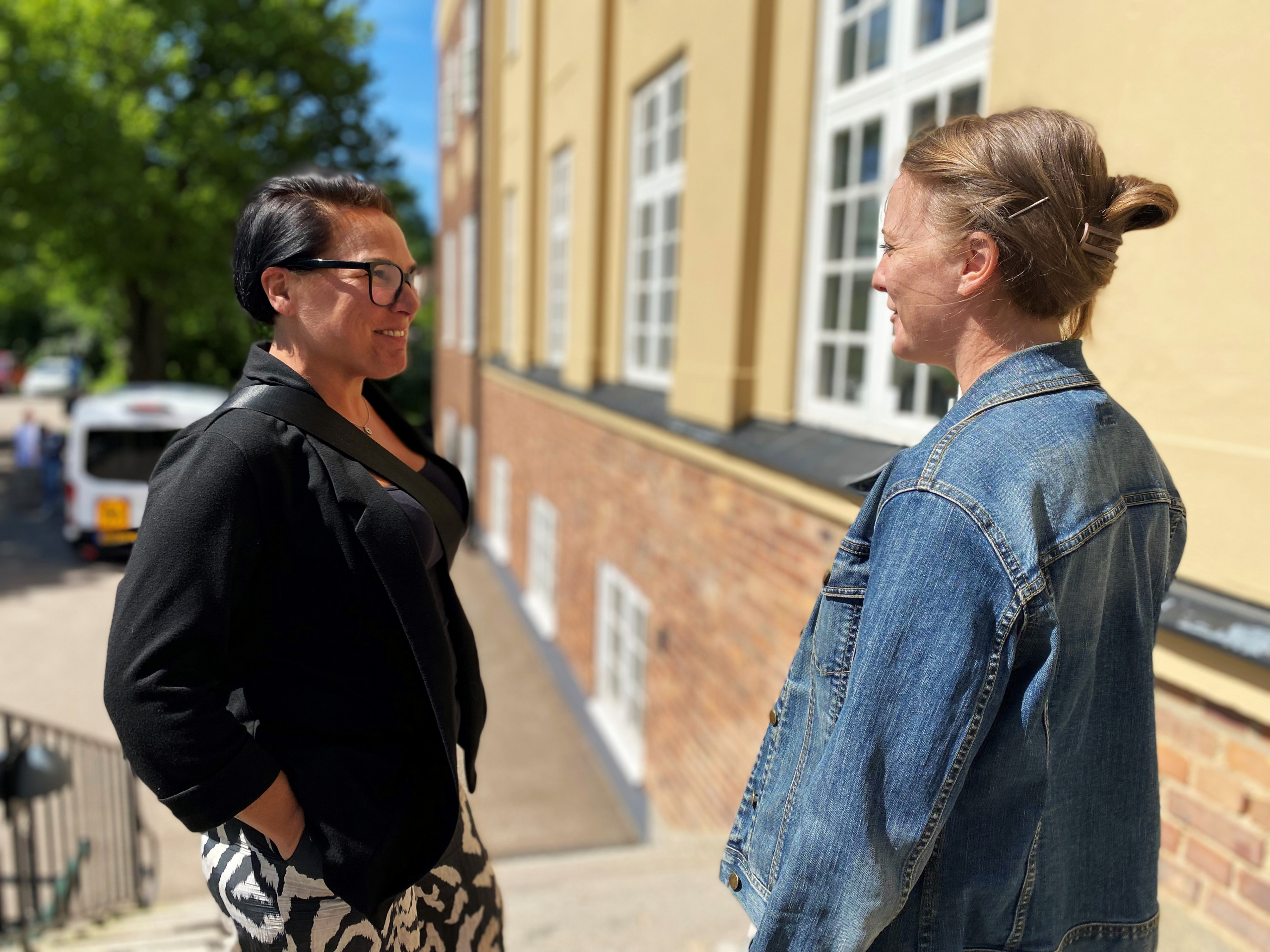 Två kvinnor står utomhus mitt emot varandra och ler