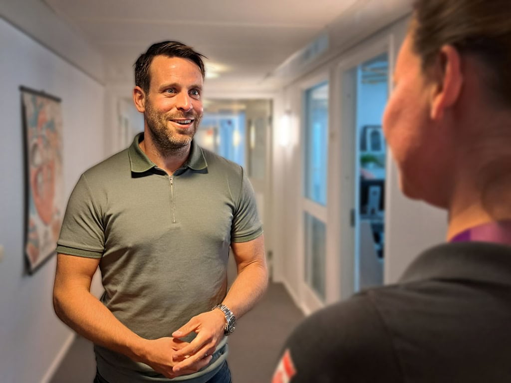 Man med kort mörkt hår och mörk skäggstubb ses stå och prata med en person som har ryggen mot kameran
