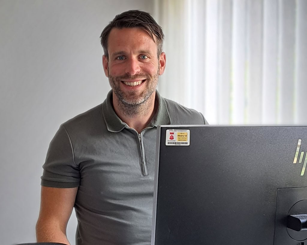 Man med kort mörkt hår och mörk skäggstubb står vid ett skrivbord bakom en datorskärm