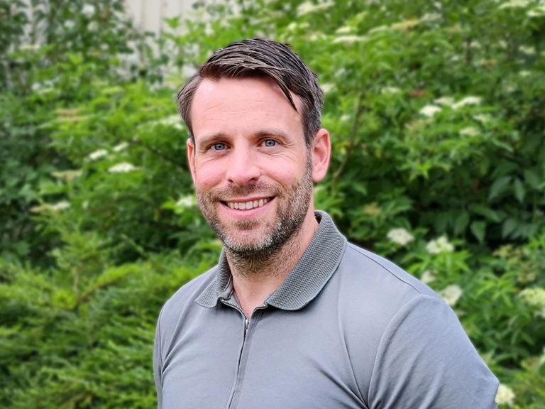 Man med kort mörkt hår och mörk skäggstubb står utomhus framför en grön buske