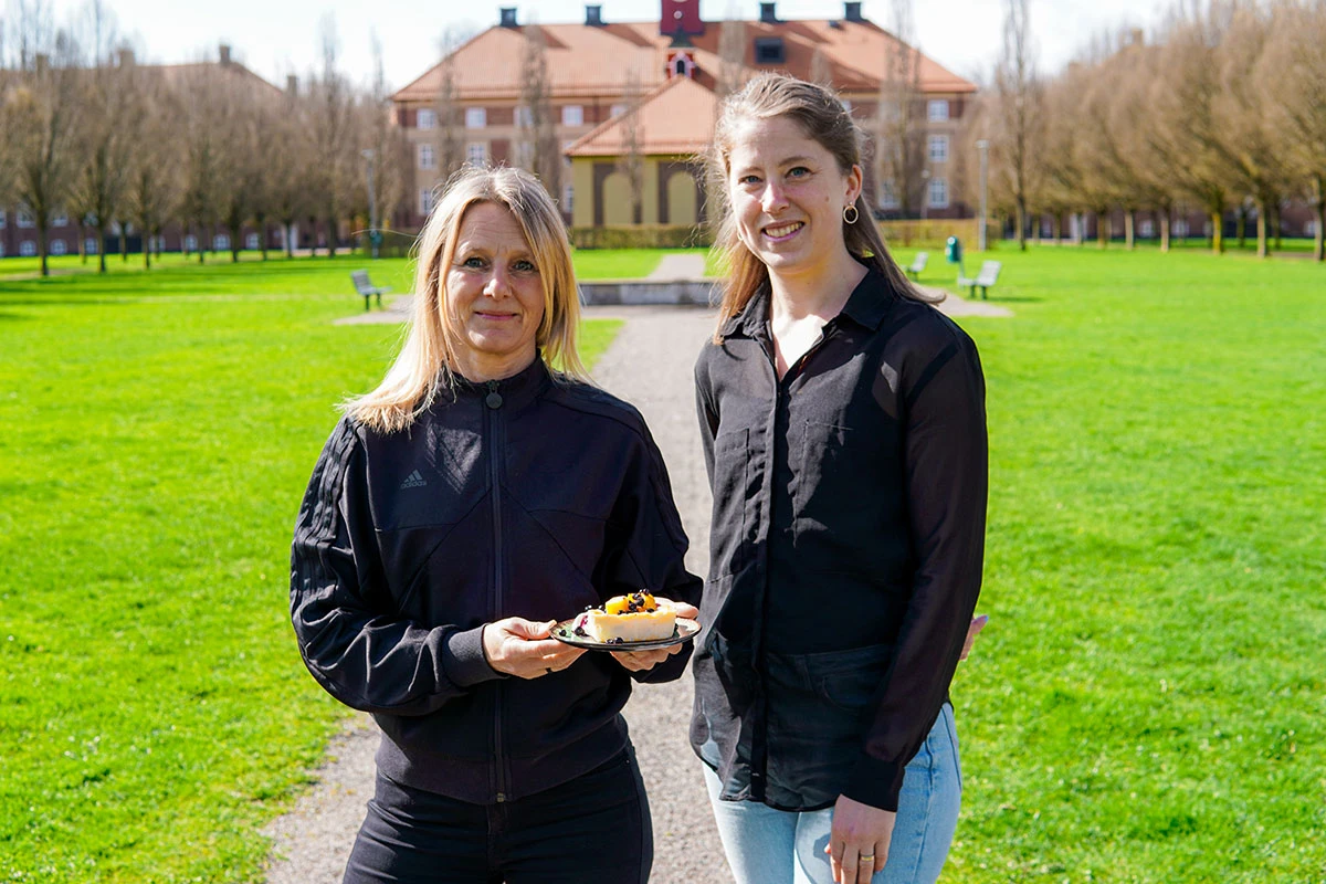 Två kvinnor står i en park vända mot kameran. Den ena håller i en tallrik med ostakaka.