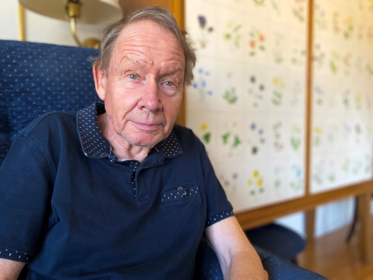 Man med blå pikétröja sitter i en fåtölj intill ett linneskåp med bilder av växter på