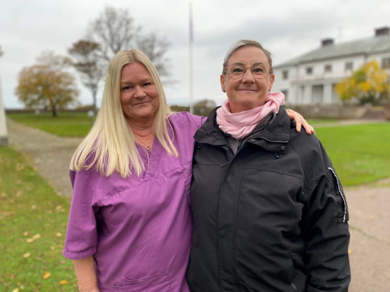 Två kvinnor står bredvid varandra utomhus i en grön lummig miljö. Den ena kvinnan håller sin arm om den andra kvinnans axlar. Båda tittar in i kameran och ler.