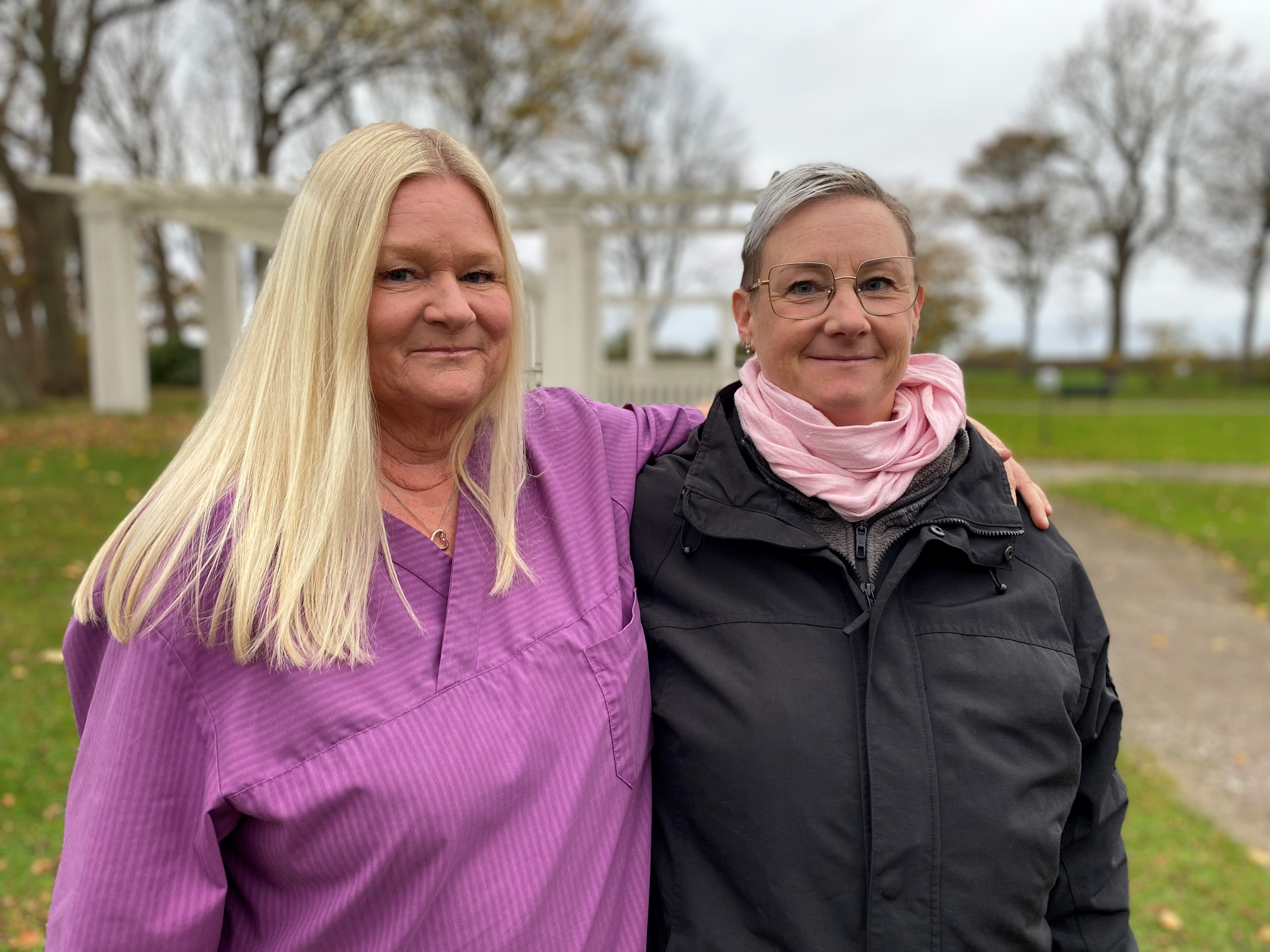 Två kvinnor står utomhus bredvid varandra. Den ena kvinnan har lagt sin arm över den andra kvinnans axlar. Båda tittar in i kameran och ler.