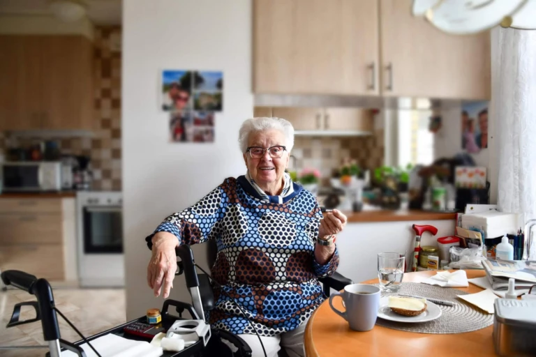 en kvinna sitter vid sitt köksbord och ser glad ut, tittar in i kameran
