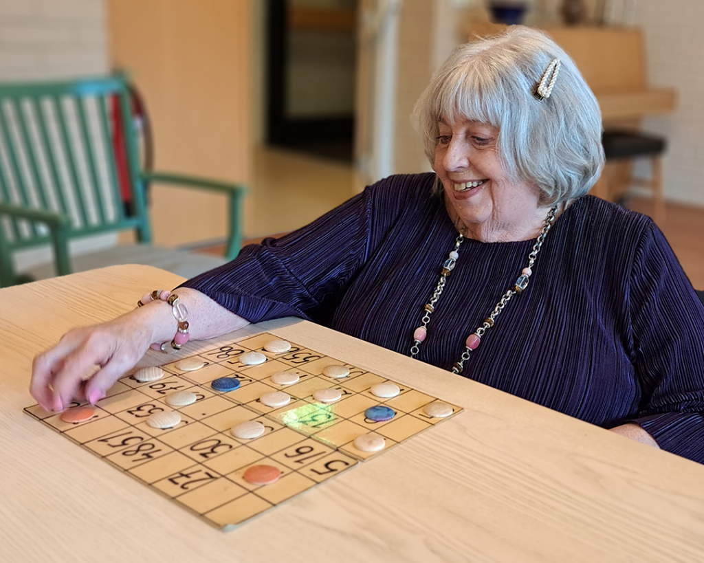 Äldre kvinna med grått hår i pagefrisyr. Mörklila blus. Spela bingo