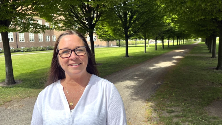 Mörkhårig kvinna i glasögon står framför en grönskande allé.