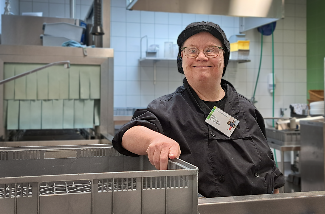 En kvinna står i ett diskrum. hon har svarta kläder, svart keps och glasögon.