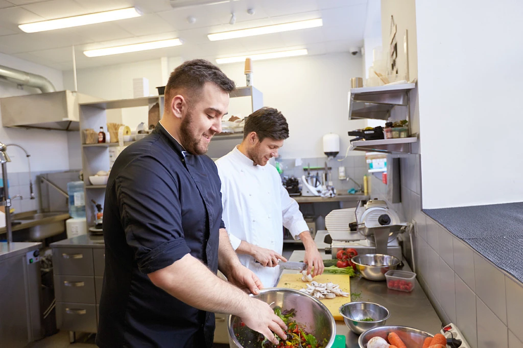 cooking food, profession and people concept - happy male chef and cook with knife chopping champignons and making salad at restaurant kitchen