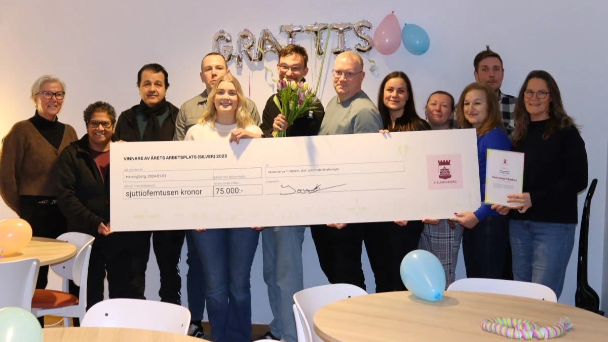 Medarbetarna som vann silver i Årets arbetsplats 2023 står och håller i en stor check. I bakgrunden hänger ballonger och det står "GRATTIS".